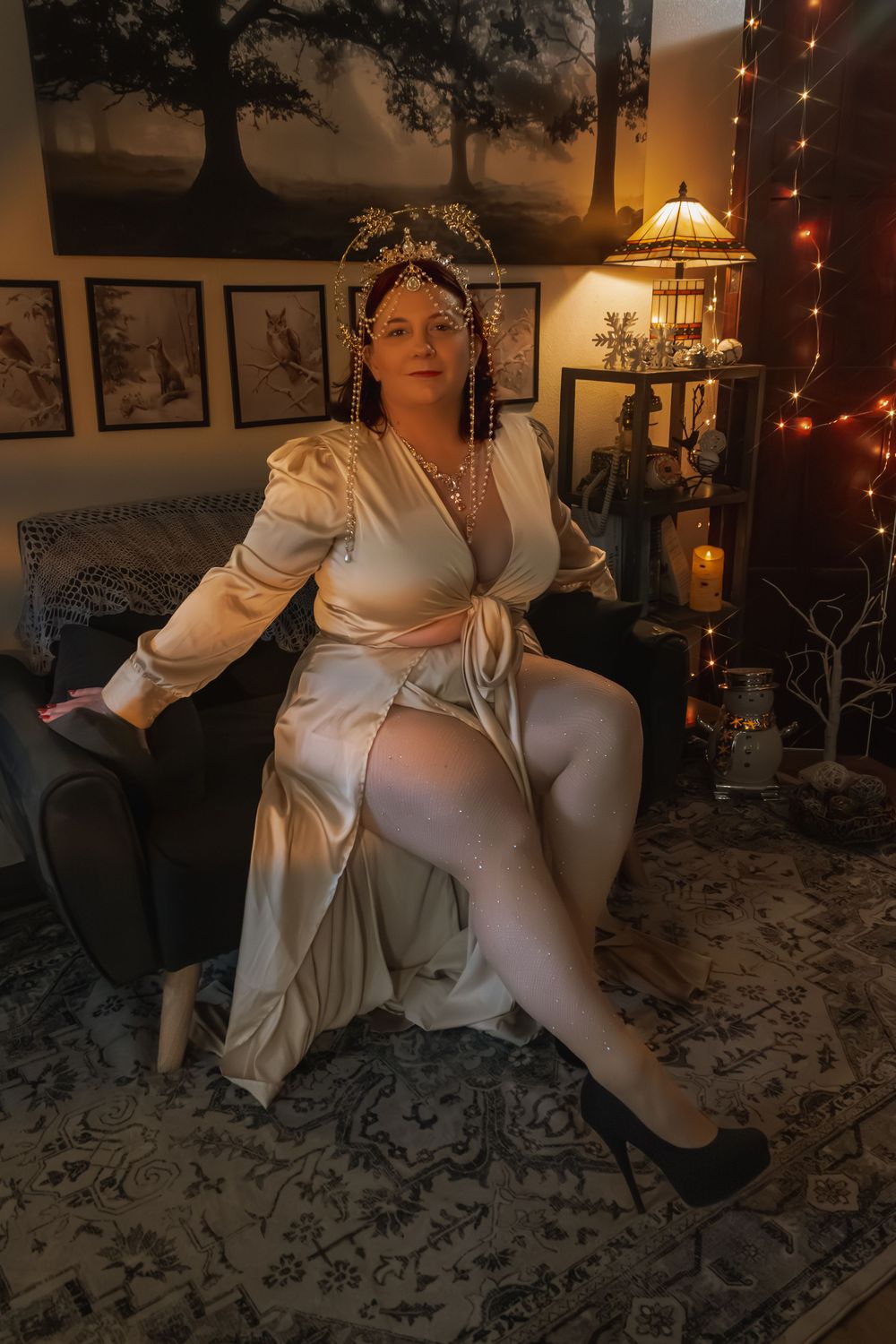 Woman in elegant cream satin dress and gold crown sits regally in decorated room with warm fairy lights.