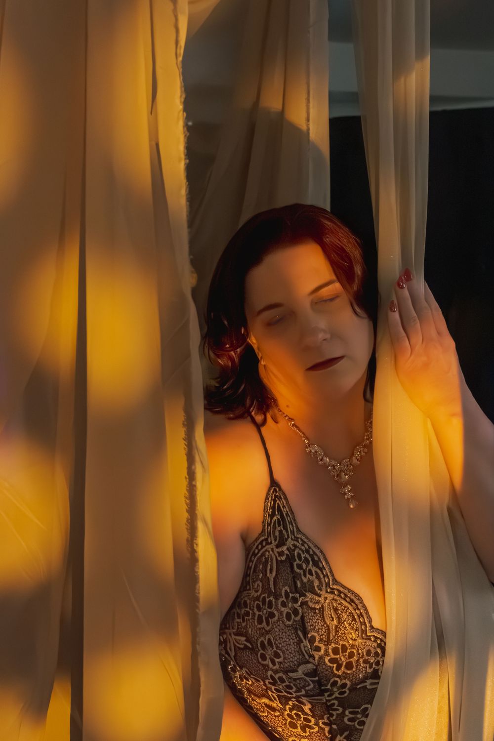 Woman in lace lingerie and necklace standing between sheer golden-lit curtains with eyes closed.