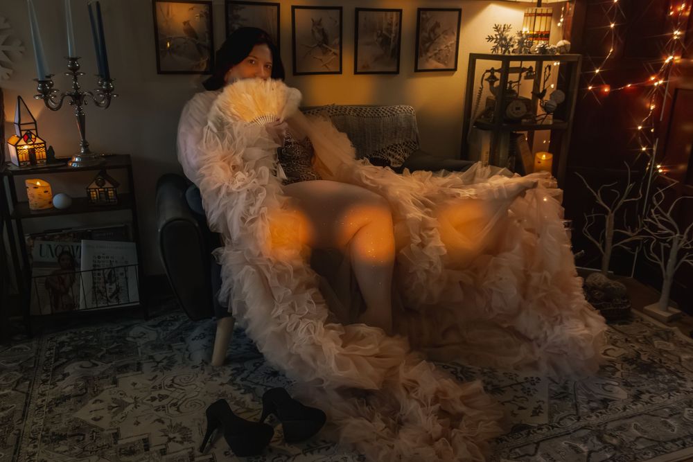 Woman in fluffy white robe relaxing in cozy dimly lit room with warm string lights and decorative shelves.