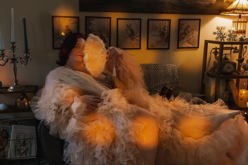 Woman in white ruffled gown holding feather fan, posed dramatically in warmly lit vintage room with candelabra.