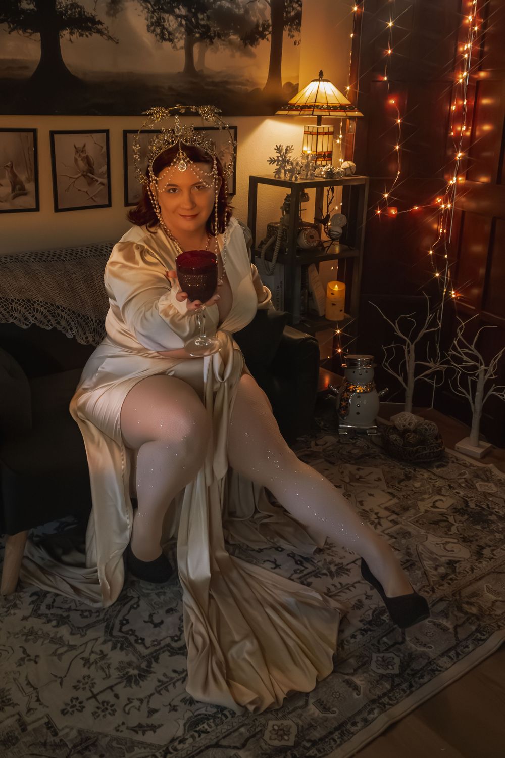 Woman in white draped gown and gold headpiece holds red wine glass in warmly lit decorated room.