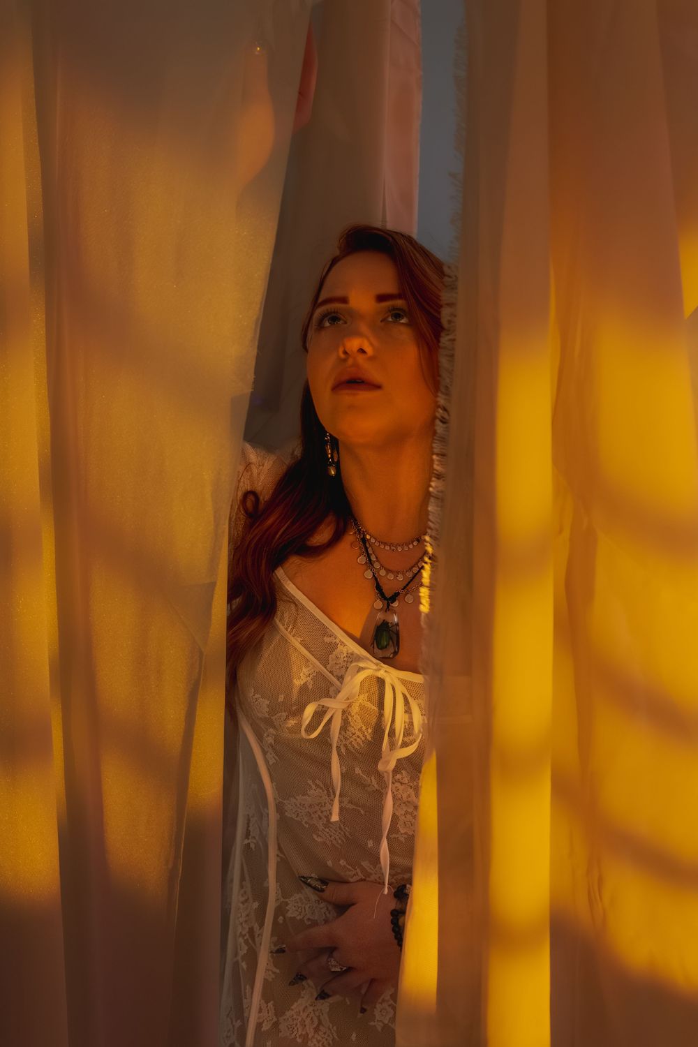 Redhead woman in white lace dress and layered necklaces peers through sheer golden-lit curtains dramatically.