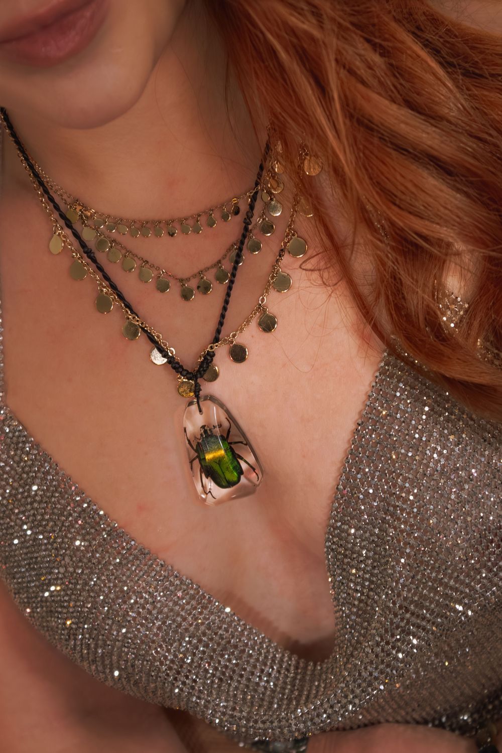 Close-up of redhead wearing layered gold coin necklace with green beetle pendant over silver glitter top.