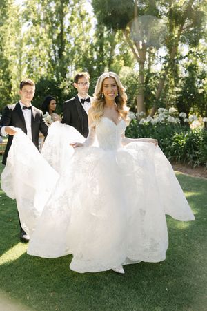 Wedding party members in white dresses and suits twirl playfully on a sunny lawn during outdoor celebration.