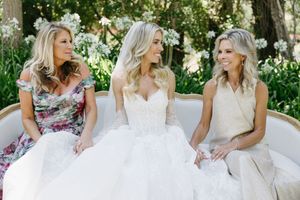 Bridal party in floral dresses and white gowns laughing together in a lush garden setting.