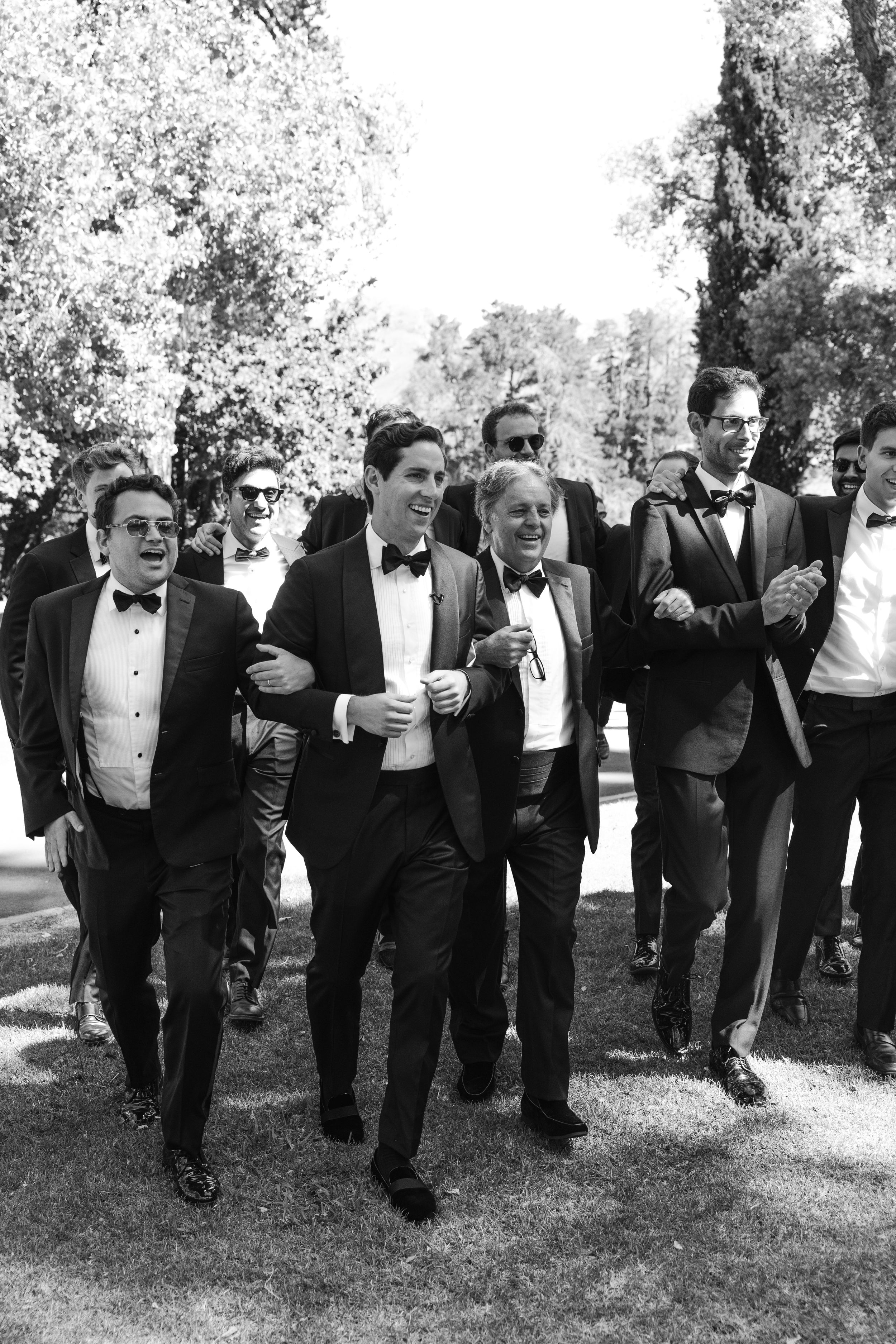 A group of groomsmen in black tuxedos walking together outdoors in a candid black and white photo.