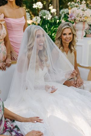 A bride in a white wedding dress and veil sits surrounded by guests at an elegant outdoor garden ceremony.