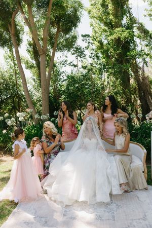 A bridal party sits together outdoors under green trees, wearing white and pink dresses in a candid moment.