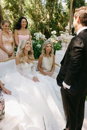 A wedding party gathers outdoors under trees, with flowing white gowns and formal attire creating an elegant scene.