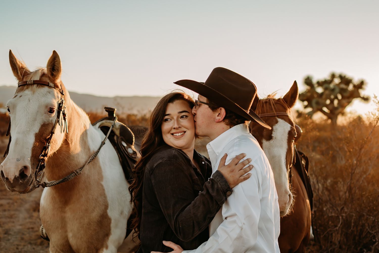 Male kissing female on cheek. They are standing between two horses with mountains, a Joshua Tree and plants behind them.