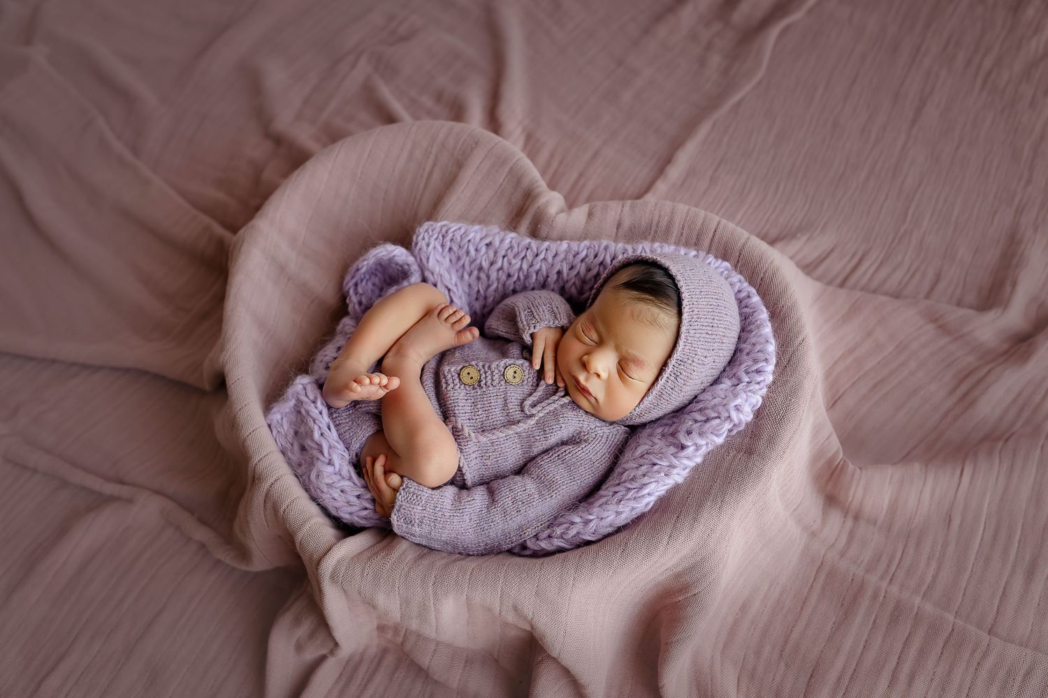 Bebe recien nacida posada dentro del prop de corazon con telas color lavanda, bebe esta durmiendo