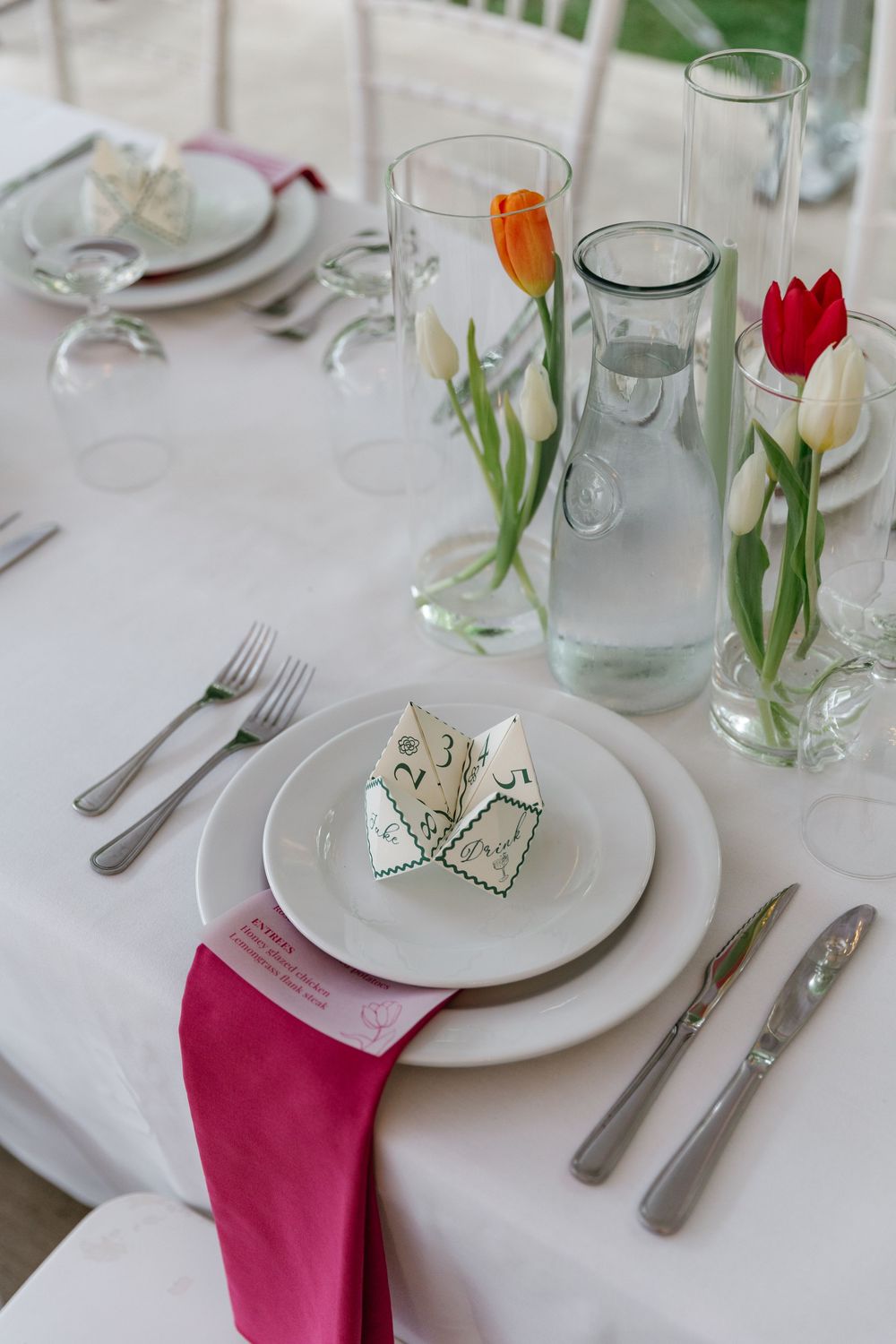 Elegant table setting with white plates, pink napkin, tulips in glass vases, and origami decor on crisp white tablecloth.
