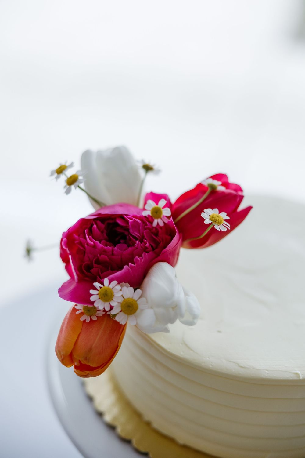 White frosted cupcake decorated with vibrant pink and orange flowers and small white daisies on a light background.