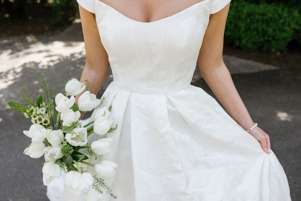 A bridal bouquet of white flowers and greenery is held against a white wedding dress in an outdoor setting.