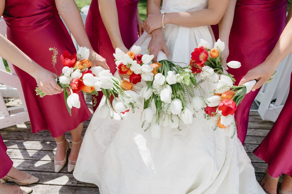 A wedding party poses outdoors wearing burgundy bridesmaid dresses and holding vibrant floral bouquets.