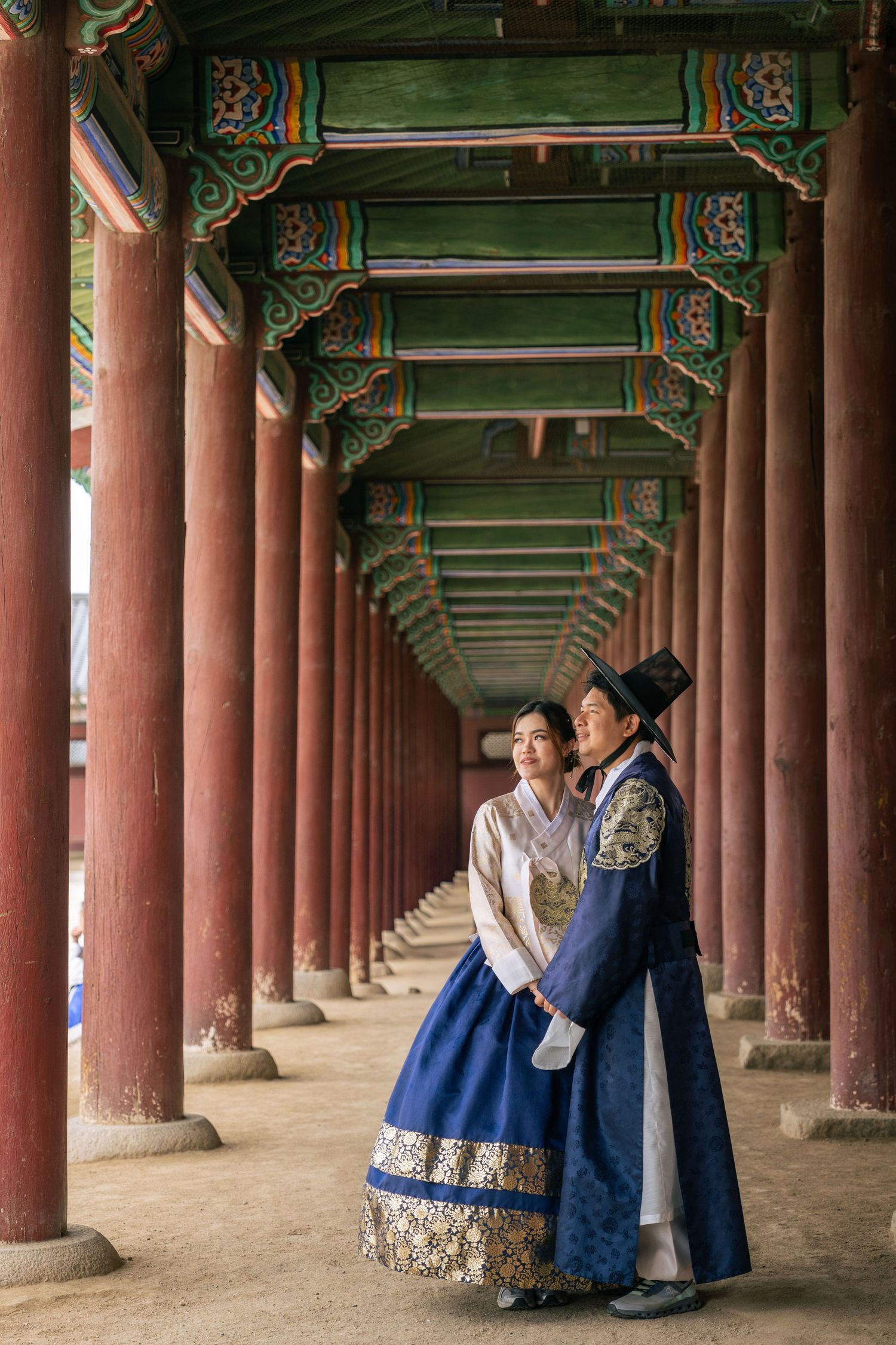 Two people in traditional Korean hanbok clothing stand in a long corridor with red pillars and ornate green ceiling at Gyeongbokgung Palace.