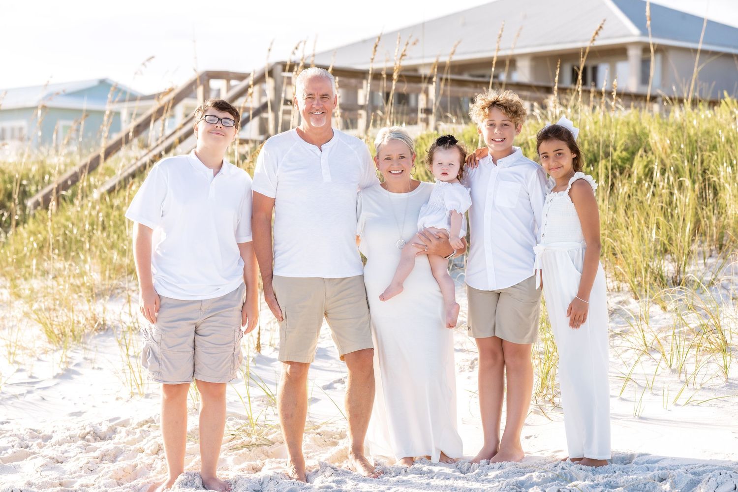 A group in white and beige outfits walks together on a sandy beach path with sea oats in the background.