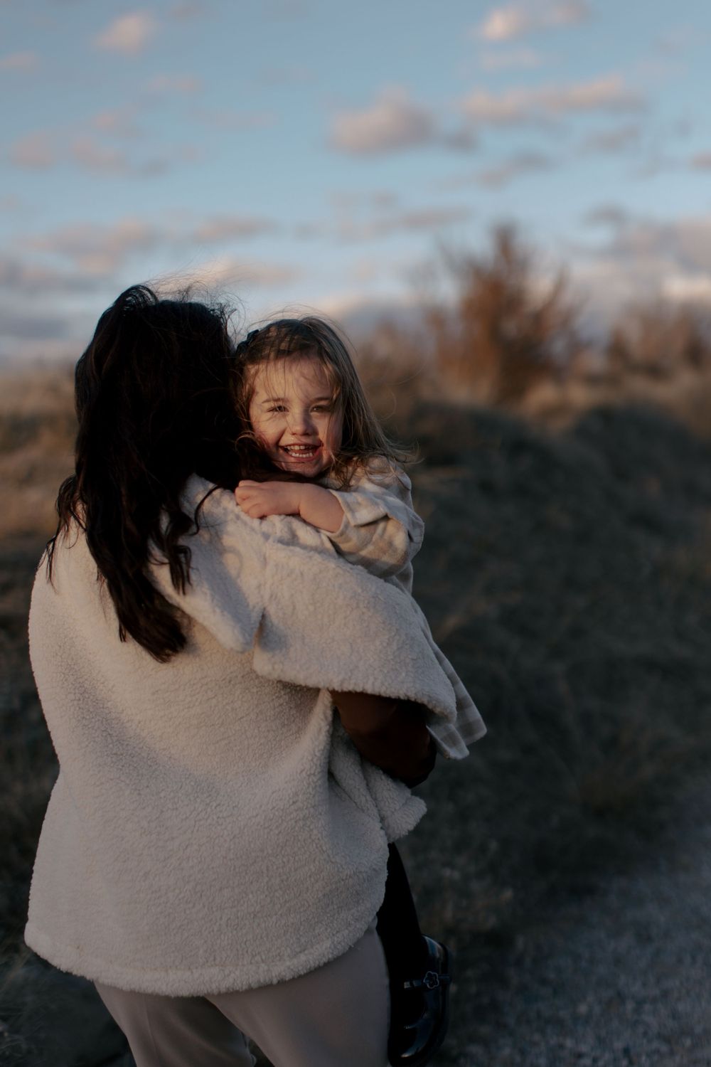A tender moment captured at sunset as a parent in a cream sweater embraces a joyful child on a hilltop.