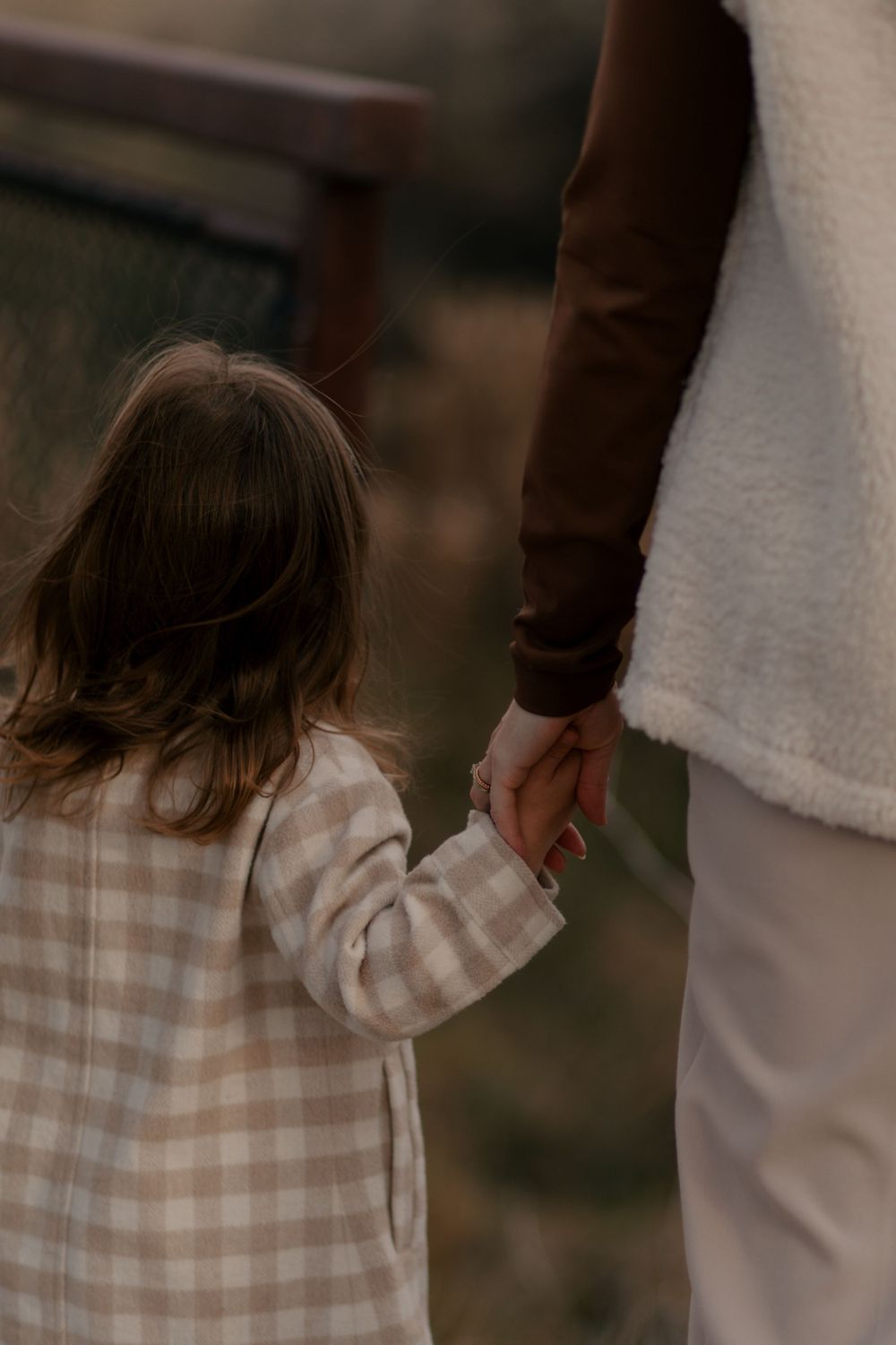 A small child in a checkered coat holds hands with an adult while walking outdoors in warm lighting.