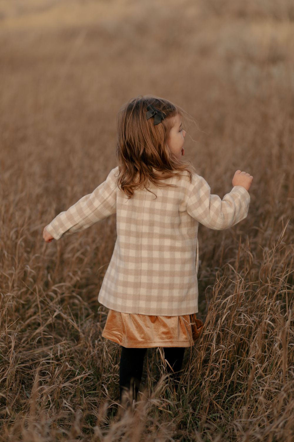 A young child in a beige checkered sweater plays in a field of tall grass during sunset.