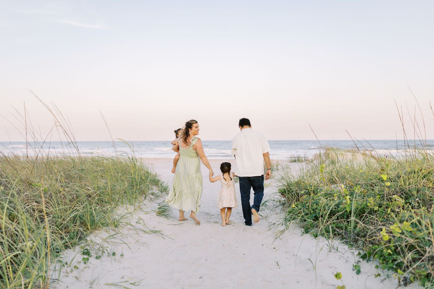 A family walks hand in hand along a sandy beach path through dunes at sunset heading towards the ocean.
