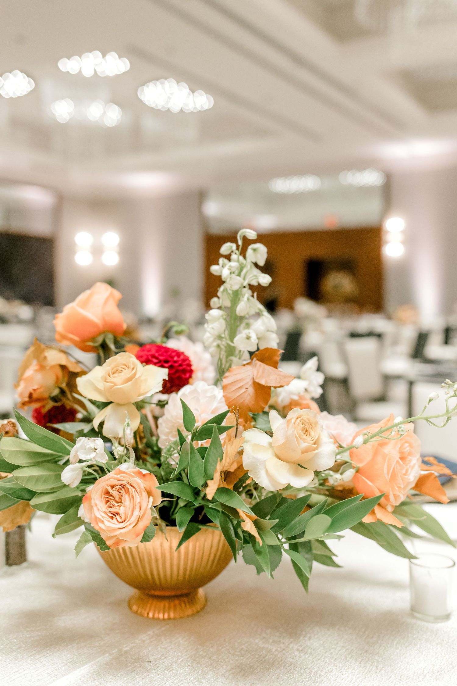 Elegant peach and coral floral centerpiece in gold vase on white tablecloth.