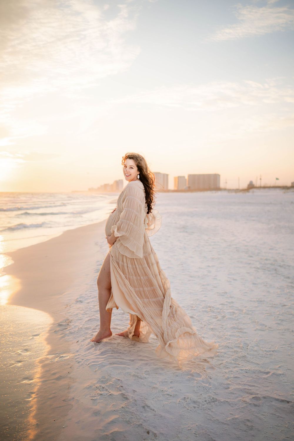 Flowy cream dress catching the wind during a sunset maternity photoshoot on a white sandy beach.