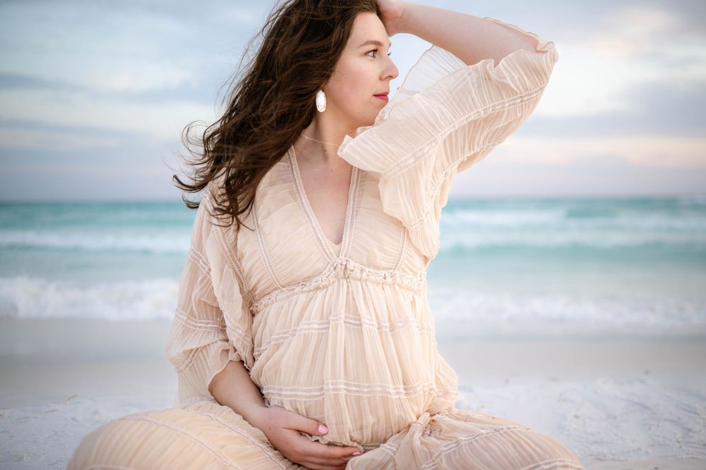 A woman in a flowing peach dress holds her pregnant belly while standing on a beach at sunset with wind in her hair.