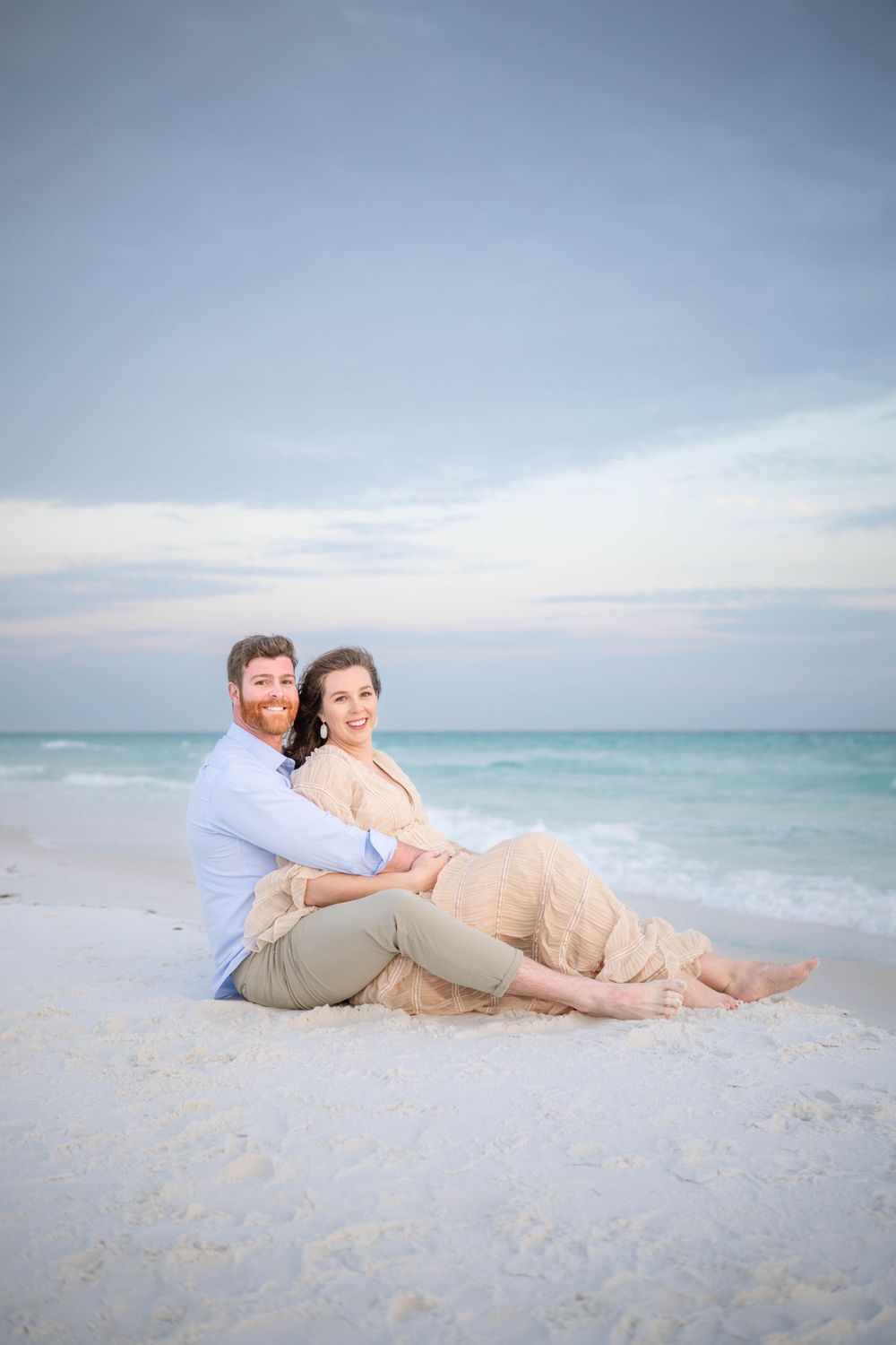 A couple sits lovingly together on a white sandy beach at sunset with turquoise ocean waves in the background.