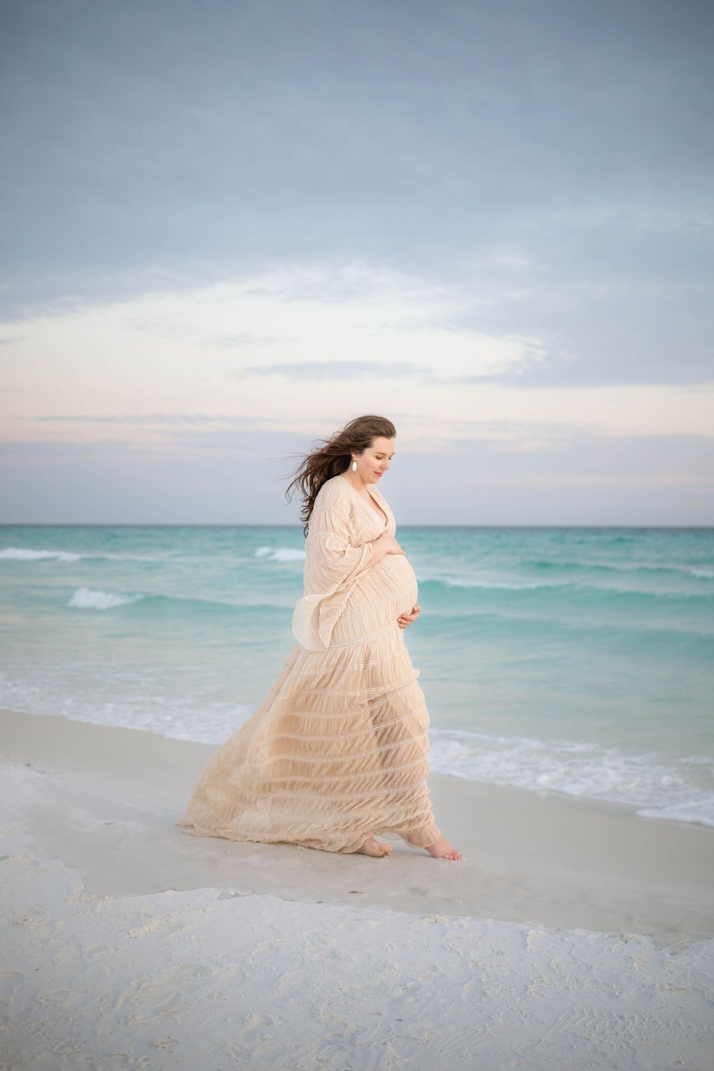 A woman in a flowing beige dress walks gracefully along a peaceful beach with turquoise waters behind her.