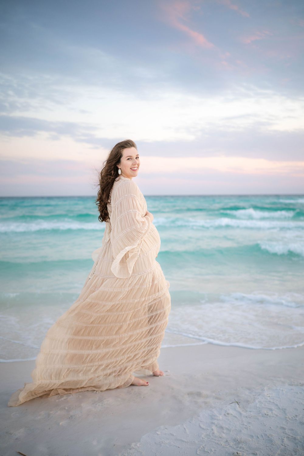 A flowing cream-colored dress catches the breeze as a woman stands on a beach at sunset with turquoise waters behind.