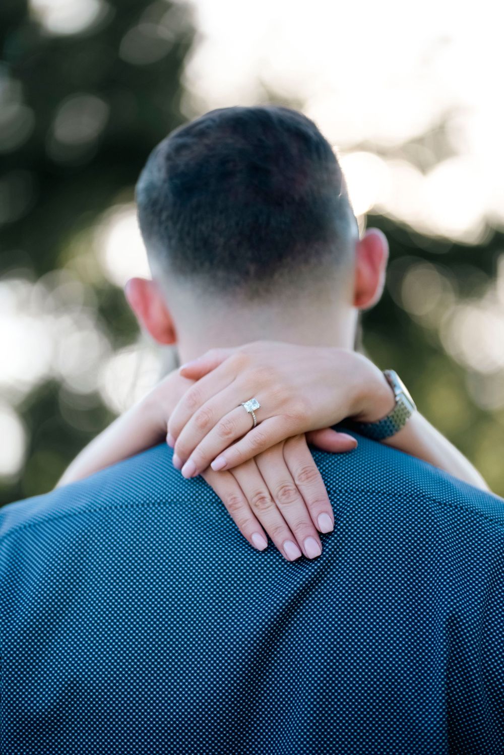 Close-up of hands displaying an engagement ring resting on a blue-clothed shoulder.