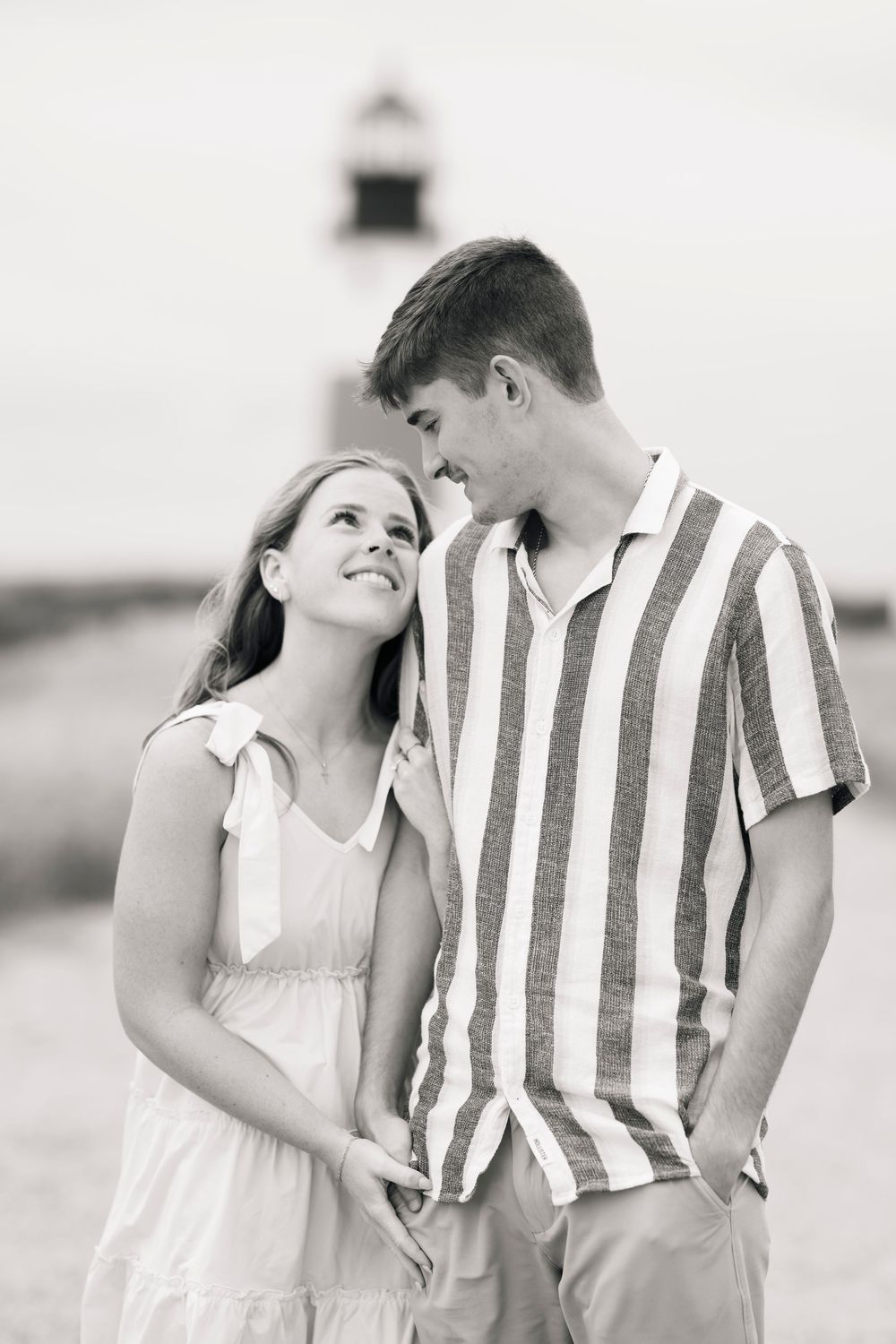A romantic black and white portrait of a couple gazing at each other with a lighthouse in the background.