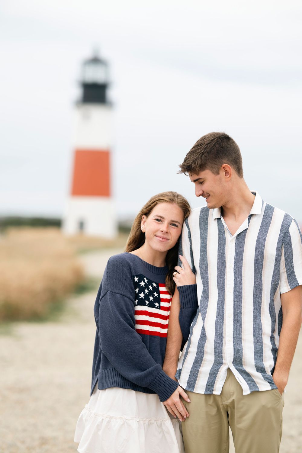 A couple poses together on a sandy beach with a lighthouse in the background.