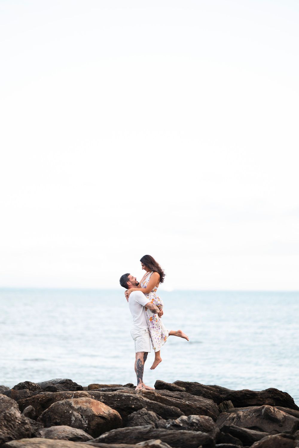 A couple embraces romantically on rocky coastal shore with ocean and white sky in background.