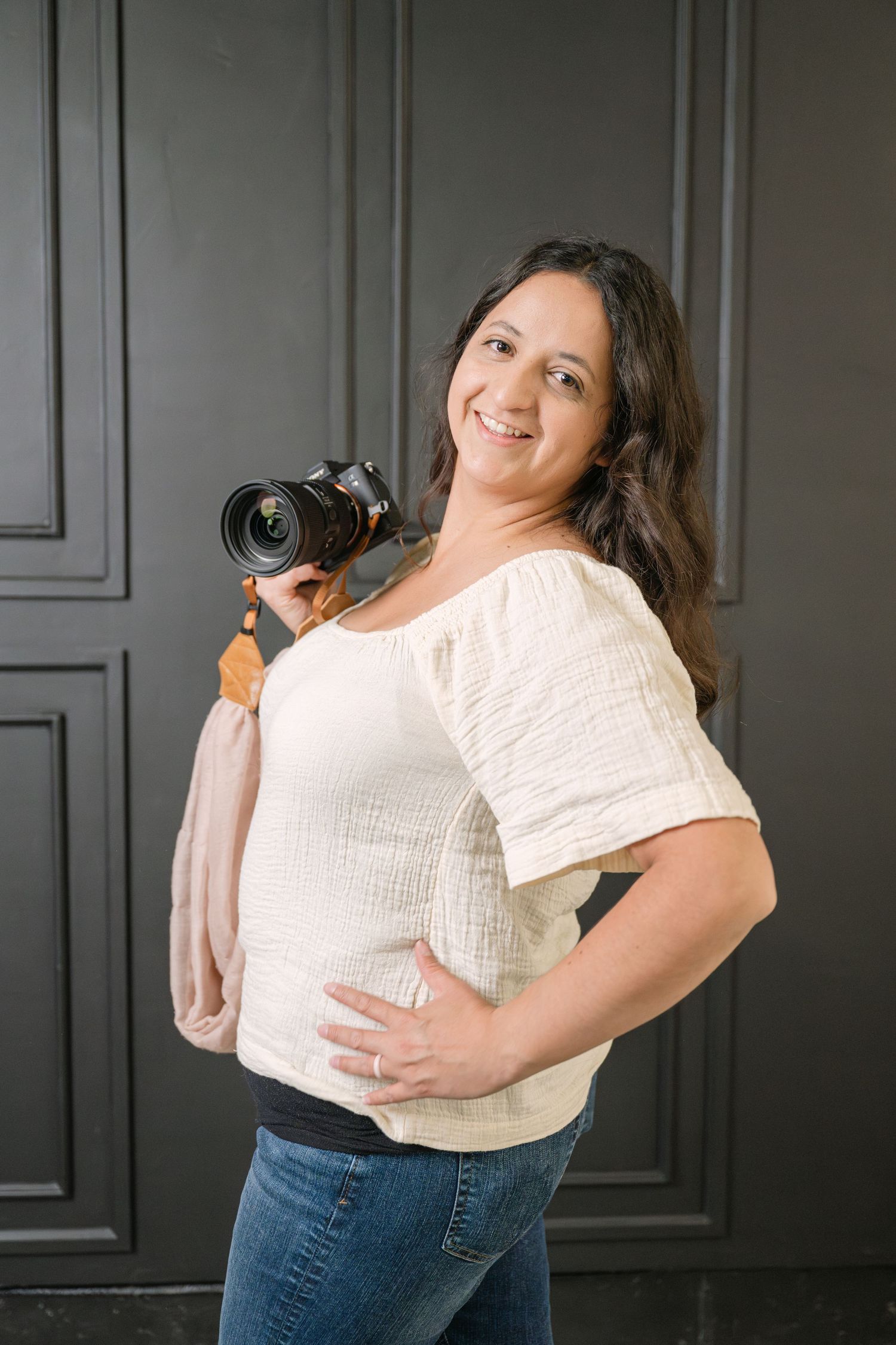 Professional photographer in white blouse and jeans smiling while holding DSLR camera against dark wall.