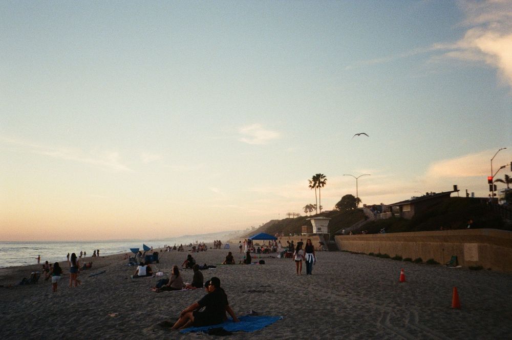 People relaxing on a sandy beach at sunset with calm ocean waves and a scenic coastal hillside in the background.