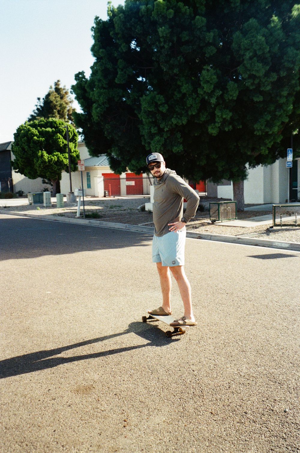 A man in light blue shorts and gray hoodie skateboarding on a sunny suburban street lined with green trees.