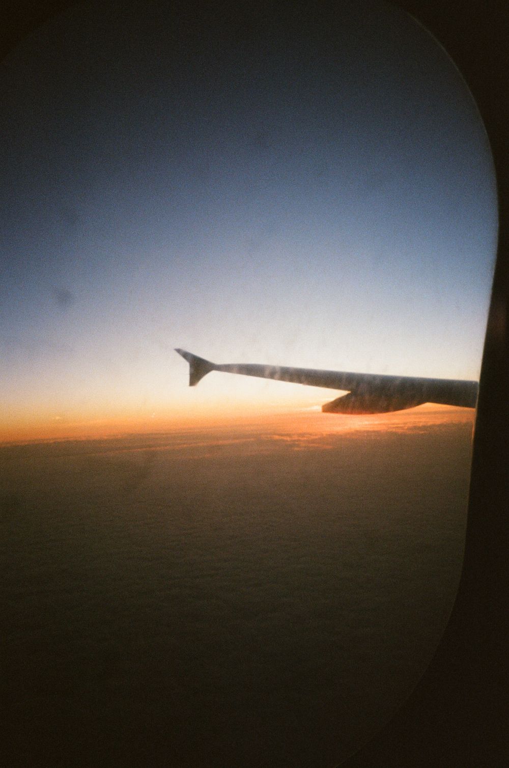 Airplane wing silhouetted against a stunning orange and blue sunset sky viewed from a passenger window at cruising altitude.