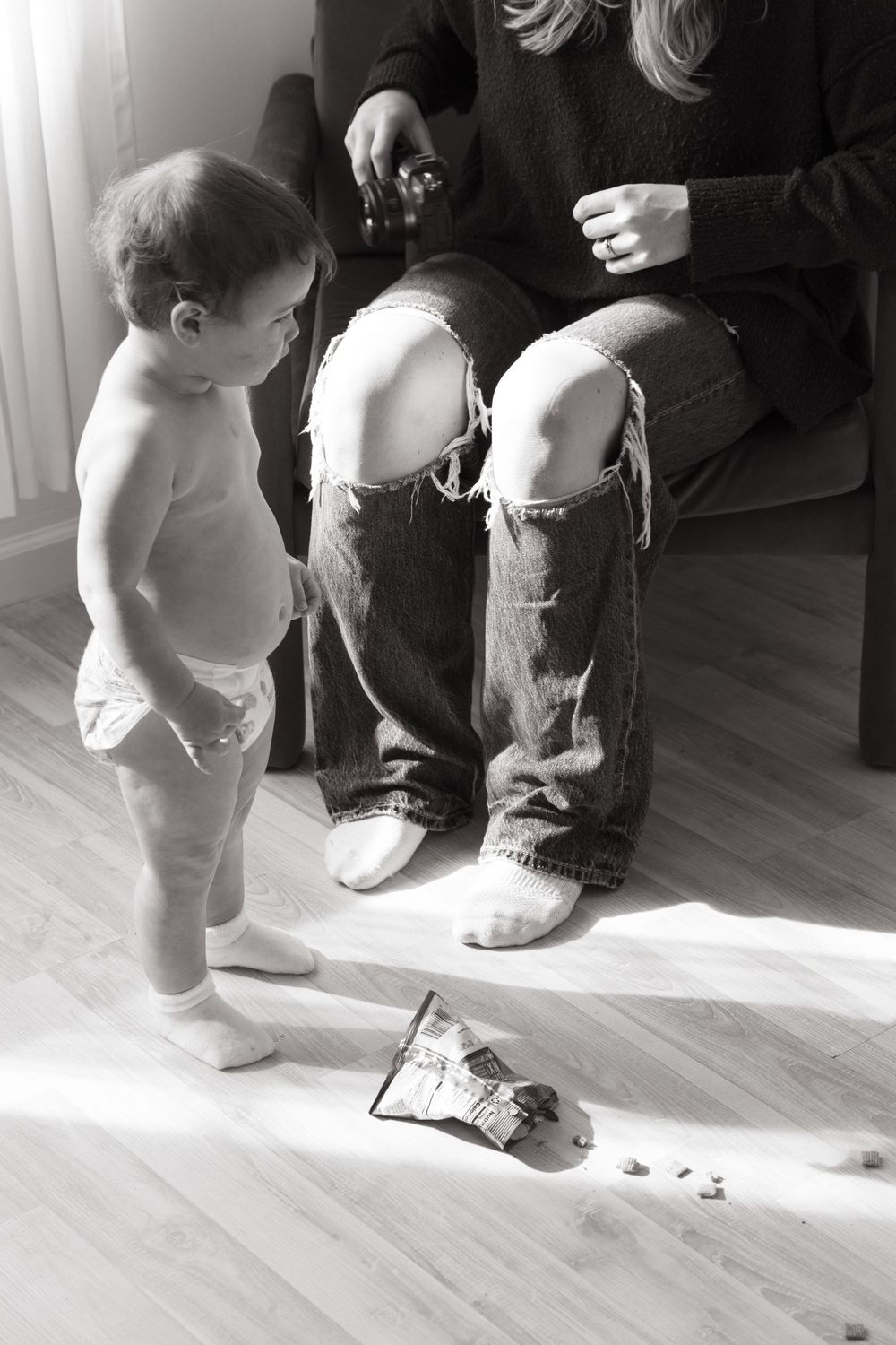 A toddler in a diaper stands beside torn jeans on a hardwood floor.