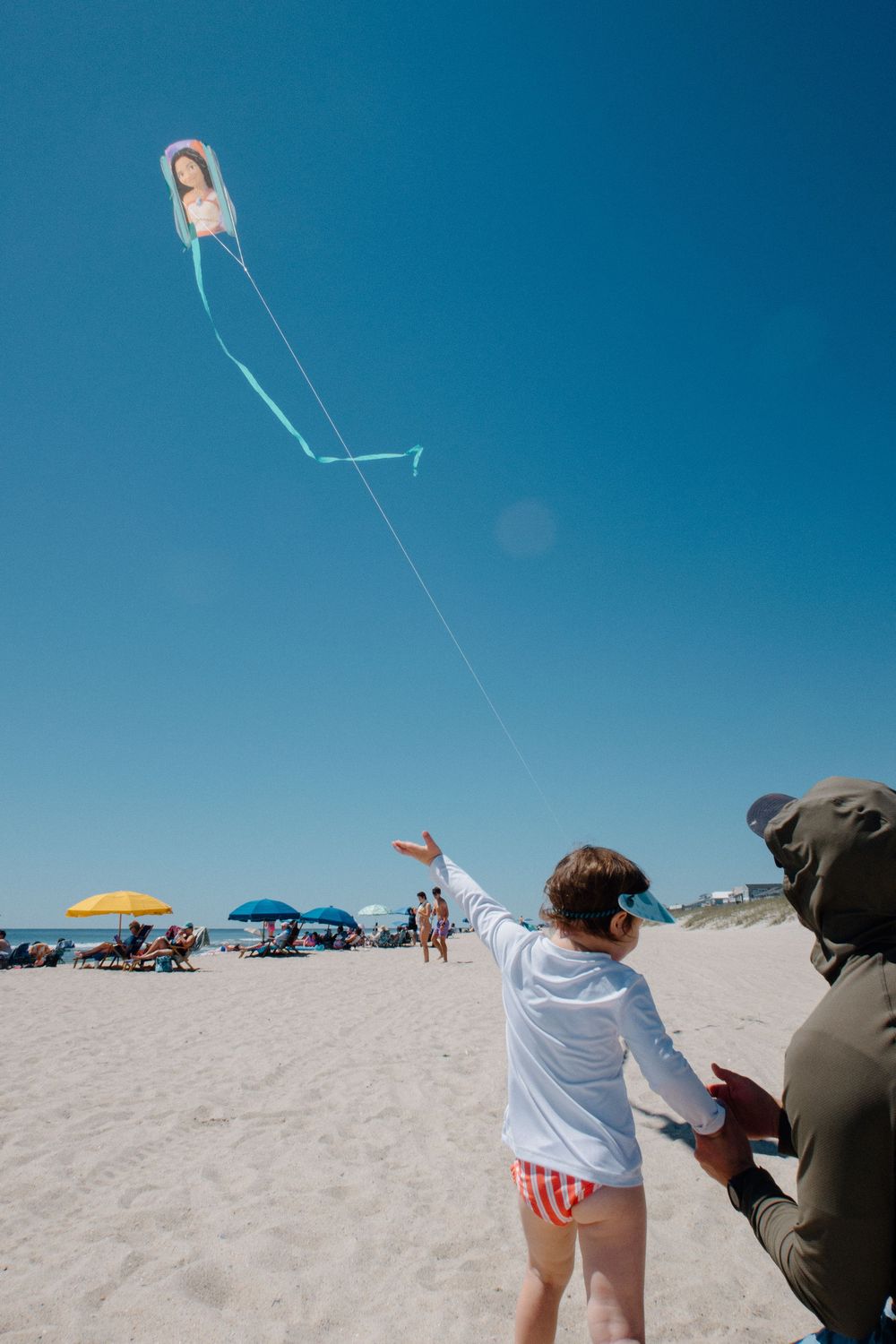 Child flying a novelty kite on a sunny beach with colorful umbrellas and clear blue sky in the background.