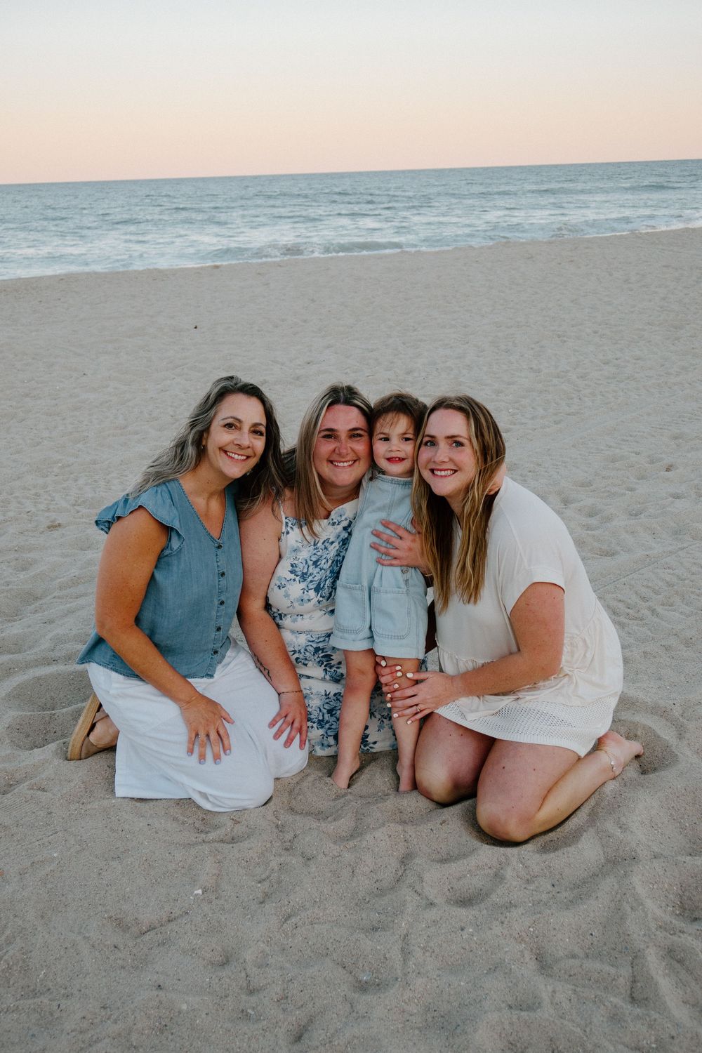 Three women and a young child smiling together on a sandy beach at sunset.