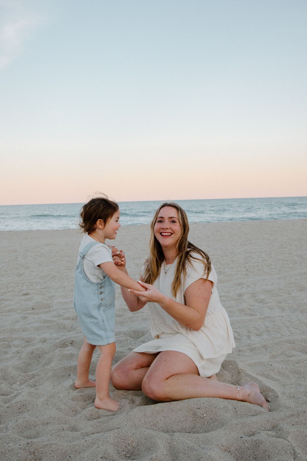 A smiling mother kneels on sandy beach holding toddler's hands at sunset, both wearing light summer dresses.