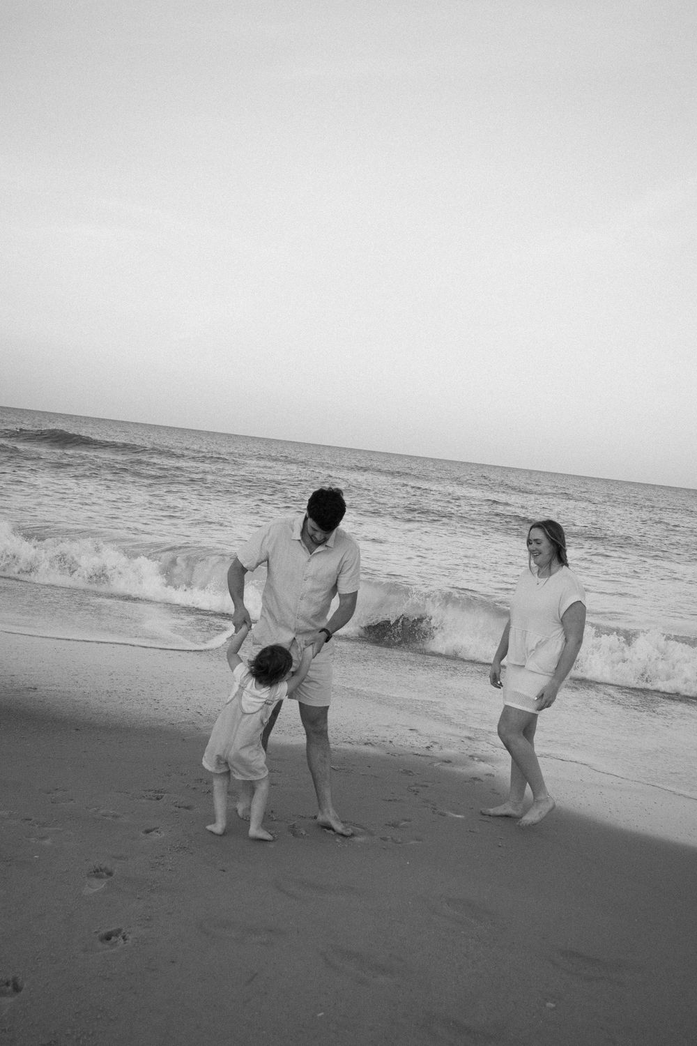 Black and white photo of a family with a toddler playing on a sandy beach with ocean waves behind them.
