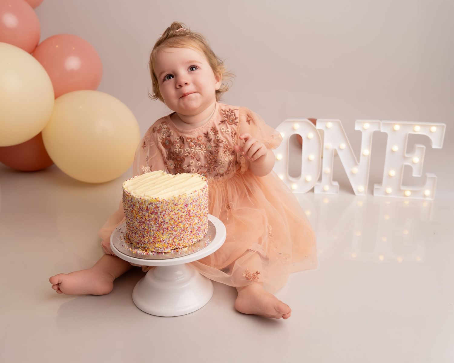 Birthday setup with pink tulle dress, sprinkle cake, and illuminated 'ONE' sign.