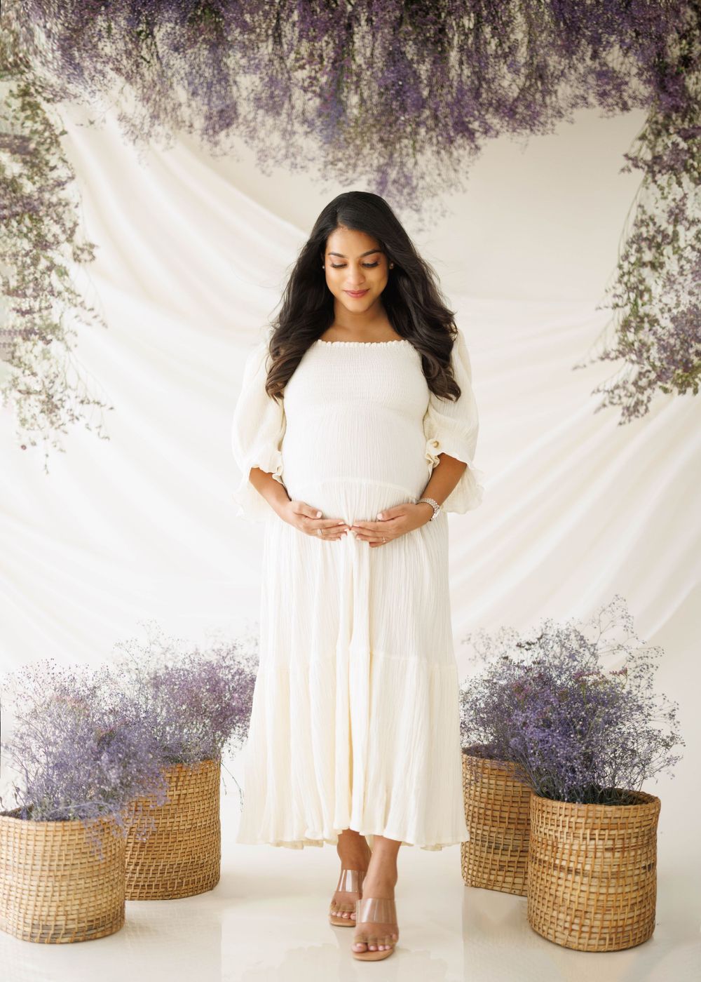 Elegant maternity photo featuring a flowing white dress against a dreamy backdrop with lavender flowers and woven baskets.