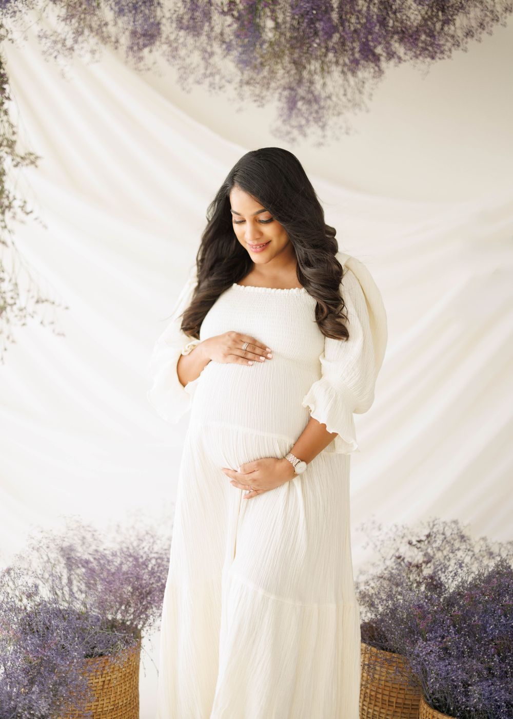 Maternity portrait with a flowing white dress and soft lighting against a white backdrop with purple floral accents.