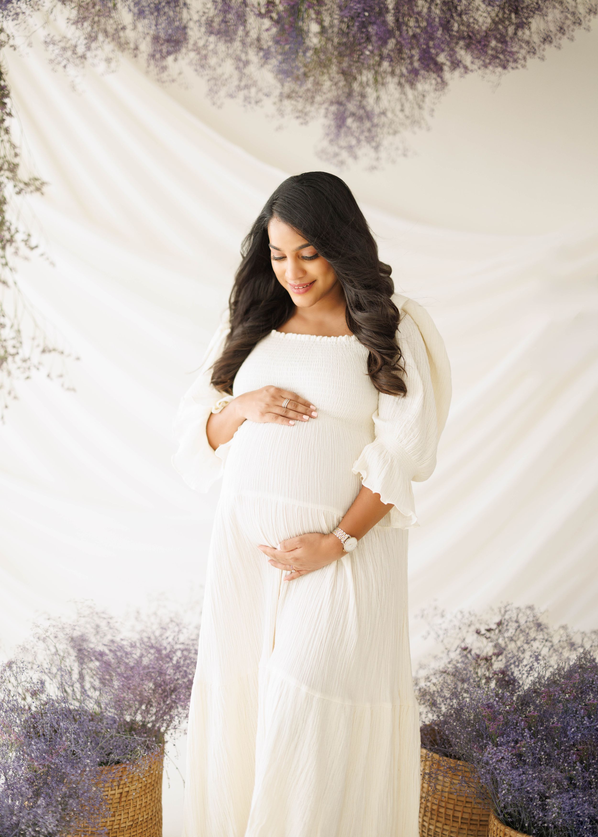 Maternity portrait with a flowing white dress and soft lighting against a white backdrop with purple floral accents.