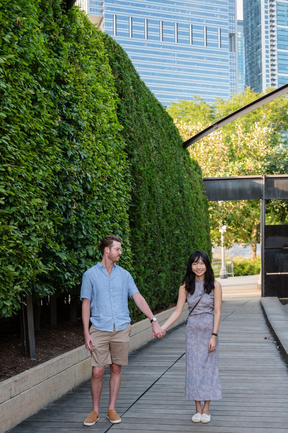 A couple holds hands while walking on a wooden deck path with lush green hedges and modern buildings in the background.