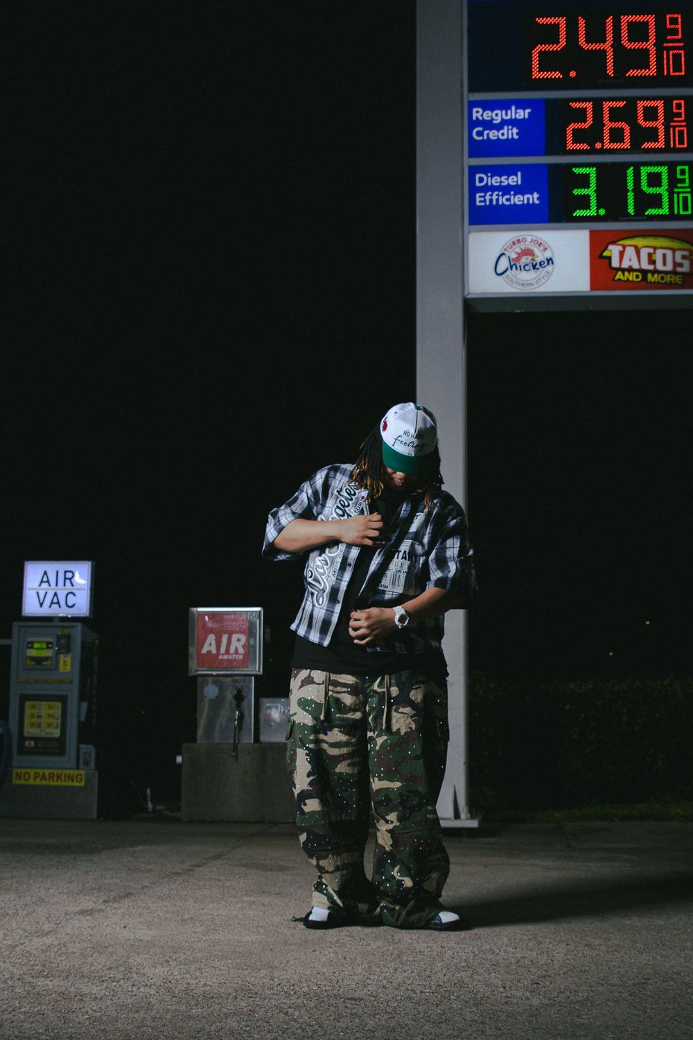 Night scene at gas station showing illuminated price display and figure in plaid shirt.